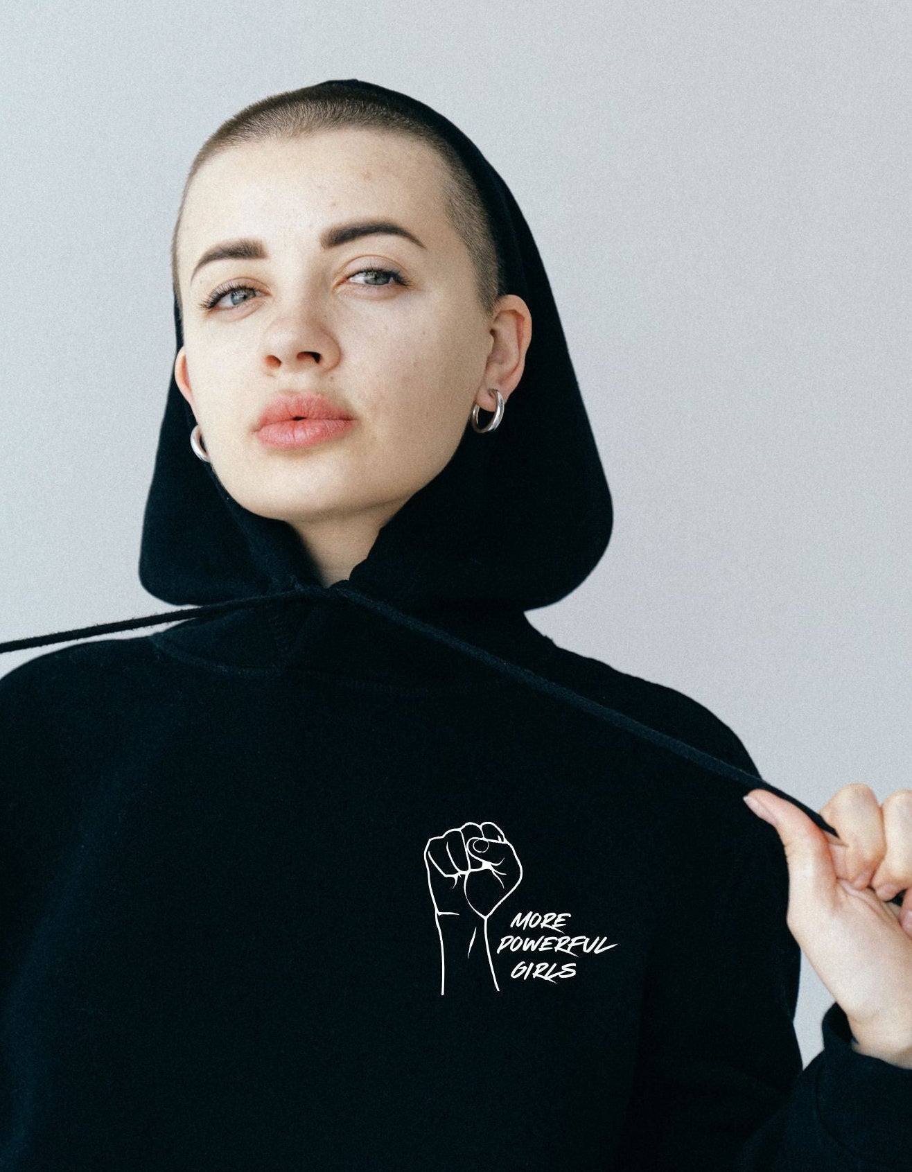Person wearing a black hoodie with a fist graphic and more powerful girls text, against a plain background