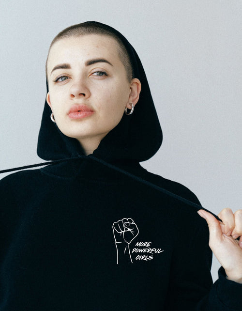 Person wearing a black hoodie with a fist graphic and more powerful girls text, against a plain background
